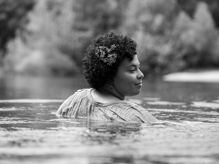 Frauenportrait im Wasser. Eine Frau mit Afro-Haar steht im Wasser, umgeben von einer natürlichen Landschaft.