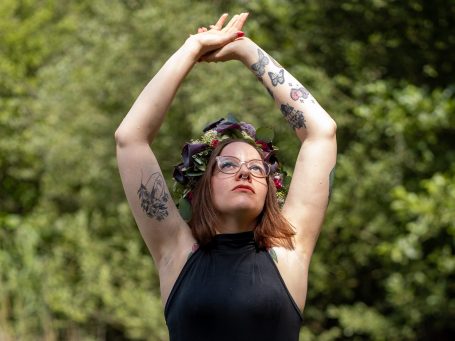 Frau mit Blumenkranz, die die Arme in die Höhe hebt, vor grünem Hintergrund.