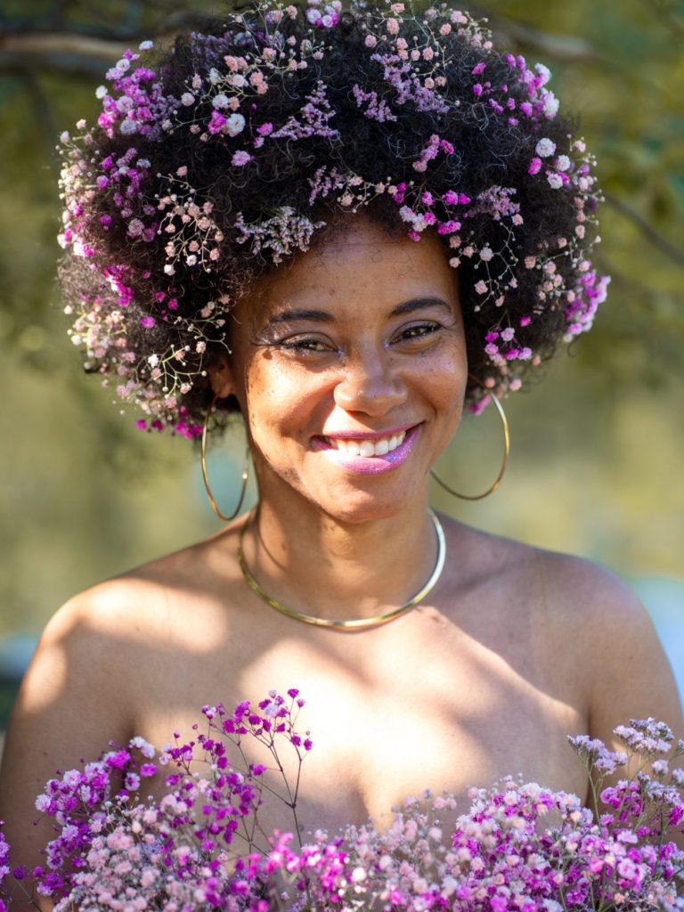 Frau mit fröhlichem Lächeln, Blumen-Afro und Blumenkranz vor natürlichem Hintergrund.