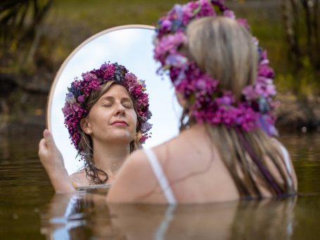 Frau mit Blumenkranz, sitzt im Wasser und blickt in einen Spiegel.
