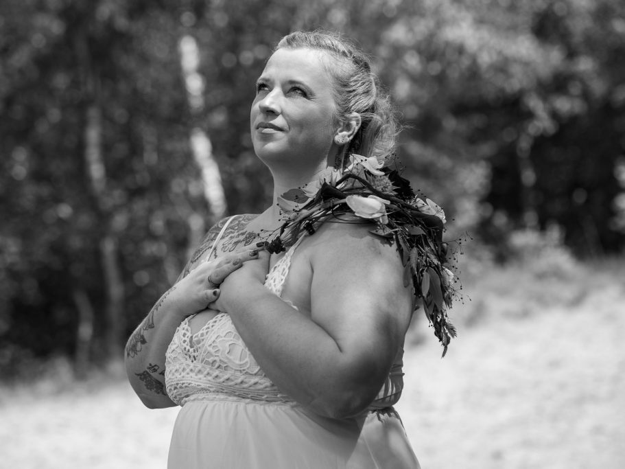 Frauenportrait- Frau in einem hellen Kleid steht lächelnd mit gefalteten Händen in der Natur.