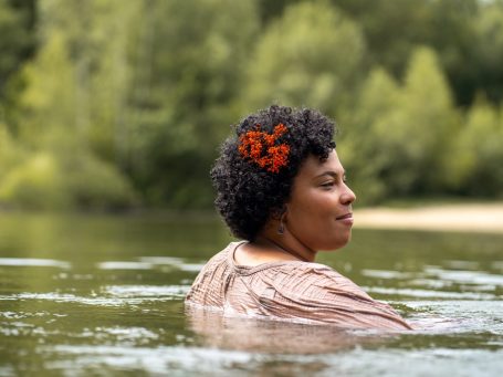Frau mit Afro-Haar und roten Blumen im Wasser, umgeben von Bäumen.